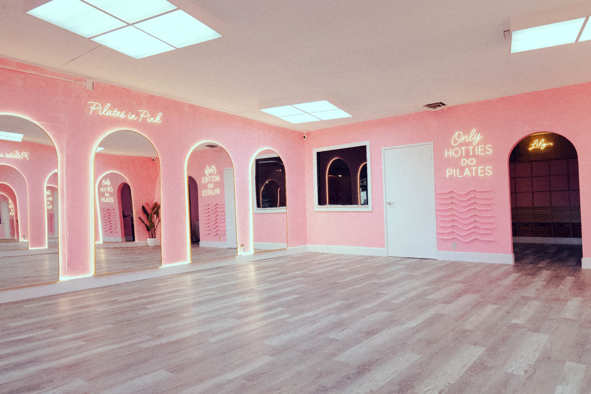 Pink studio room with arched lit mirrors, light wood floors and neon signs reading Pilates in Pink and Only Hotties Do Pilates