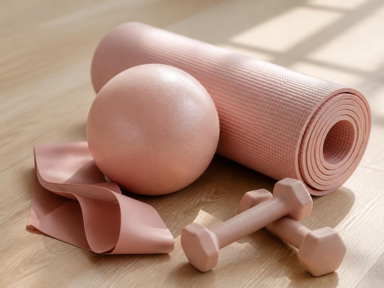 Rolled pink exercise mat with a small ball, resistance band and light hand weights on a wood floor